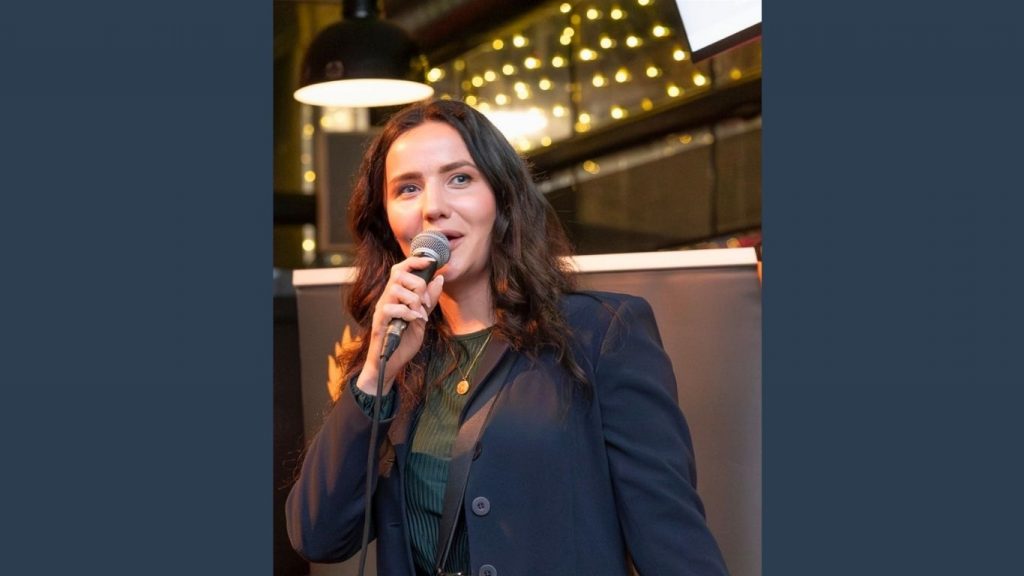 Franziska Barth hält einen Experten-Vortrag zur betrieblichen Gesundheitsförderung (BGF) im Business-Outfit mit Mikrofon auf einer Event-Bühne.